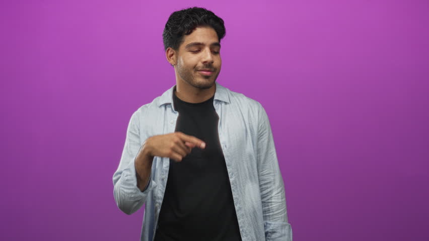 Man points finger upward in studio against purple backdrop, wearing open light blue shirt over black tshirt; curiosity pondering.
