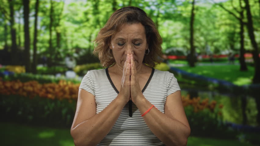 Woman standing with hands pressed together in forest setting under bright green tree canopy; serenity.