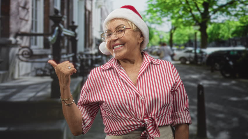 Woman wearing santa hat and glasses points thumb over shoulder on a leafy city street while smiling in a striped tied shirt and beige trousers; holiday joy.