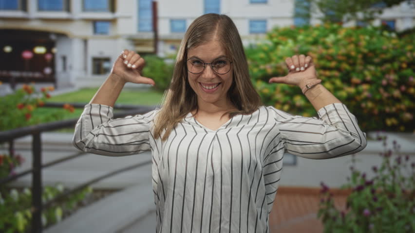 Woman pointing thumb to building facade among garden flowers outdoors with smiling grin and raised arms; confidence.