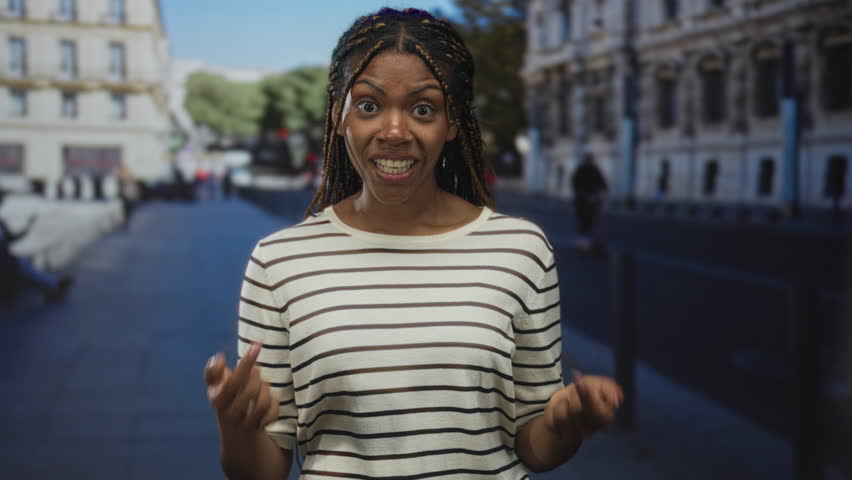 Woman points fingers to chest on street in front of building wearing striped shirt and braided hair; surprise reaction.