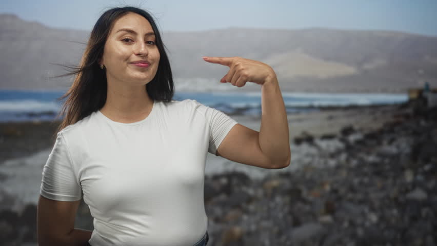 Young woman points finger to chest on rocky seaside shore wearing tshirt with windblown hair while standing outdoors; confidence empowerment.