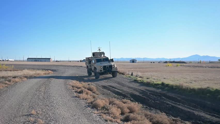 A military tactical vehicle drives on a rocky road against a backdrop of vast plains and mountains. This type of vehicle is designed to handle a variety of terrain conditions.