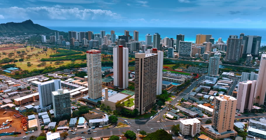 Approaching few high-rises over the low-rise houses. Modern cityscape of Honolulu, Hawaii, USA. Waikiki scenery at backdrop. Aerial view.