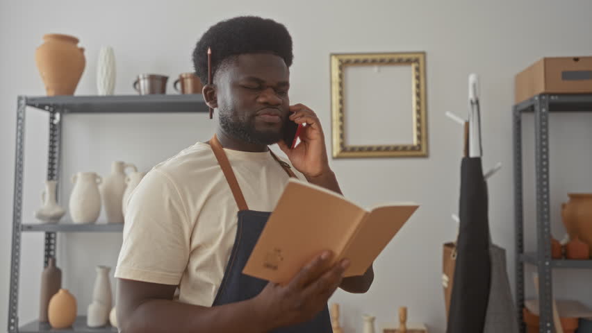 Man in denim apron talking on smartphone while holding notebook with pencil tucked behind ear in studio; focused craft.