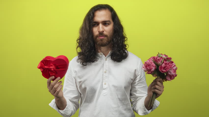 Man holding red heart box and pink bouquet in lime green studio, puffing cheeks and smiling with closed eyes; playful romance.