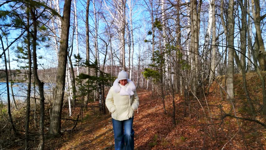 Woman in yellow jacket walks along a scenic forest path, surrounded by trees and snow, as the camera follows with a smooth dolly movement capturing the serene atmosphere