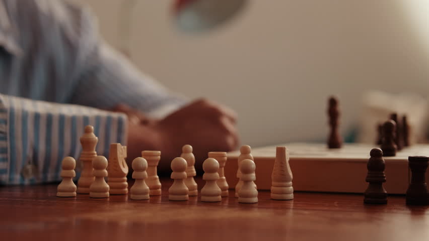 A person sets up white chess pieces on a wooden table. The black chess pieces are already set up on the board in the background.