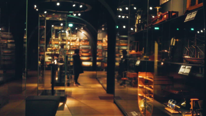 Museum gallery showcasing blurred warship models with reflected lights and a woman observing the exhibits in an artistic setting.