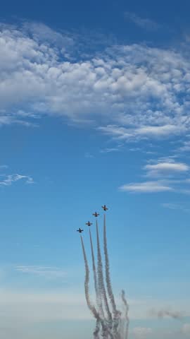 Four jets ascend in close formation leaving smoky trails behind, while one aircraft separates from the group, diverging to the right against blue sky. Vertical.
