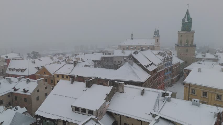 Aerial drone flyover above snowy rooftops in Lublin, Poland, revealing historic church towers and old architectural landmarks covered in fresh white snow, creating a peaceful winter cityscape.