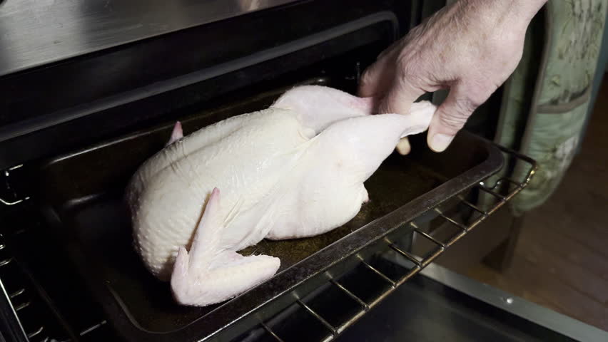 Closeup of a man’s hand slowly placing a raw uncooked whole chicken into a kitchen oven tray, ready for roasting.