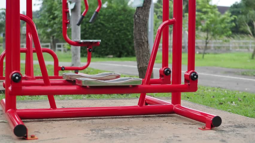  Man wearing black pants, a green shirt and green socks exercises outdoors with a red stainless steel exercise machine in a park.