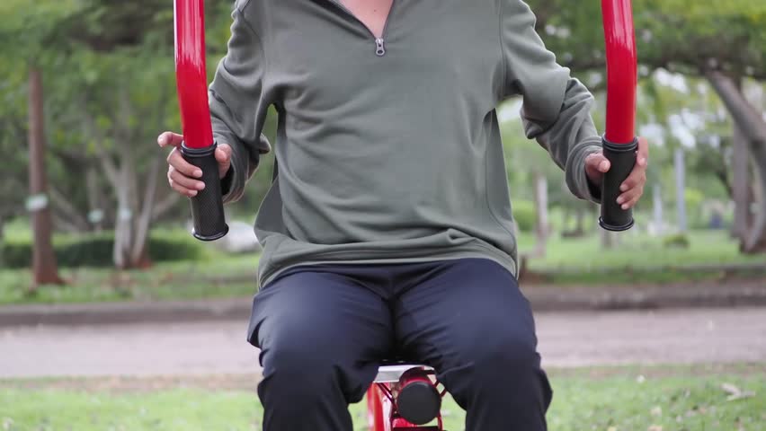  Man wearing black pants, a green shirt and green socks exercises outdoors with a red stainless steel exercise machine in a park.