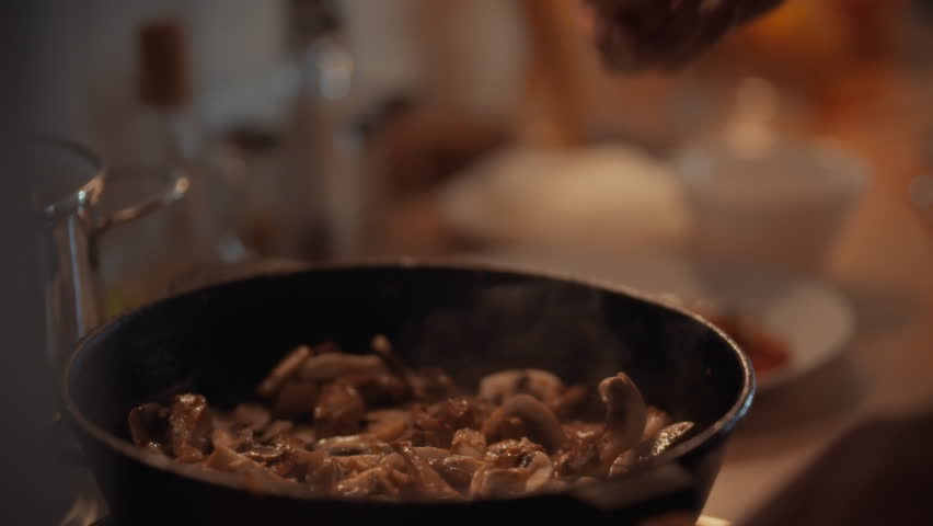 A hand adds salt to a skillet of cooking champignon mushrooms, creating a savory flavor in a warm kitchen. The action takes place during meal preparation, highlighting the cooking process
