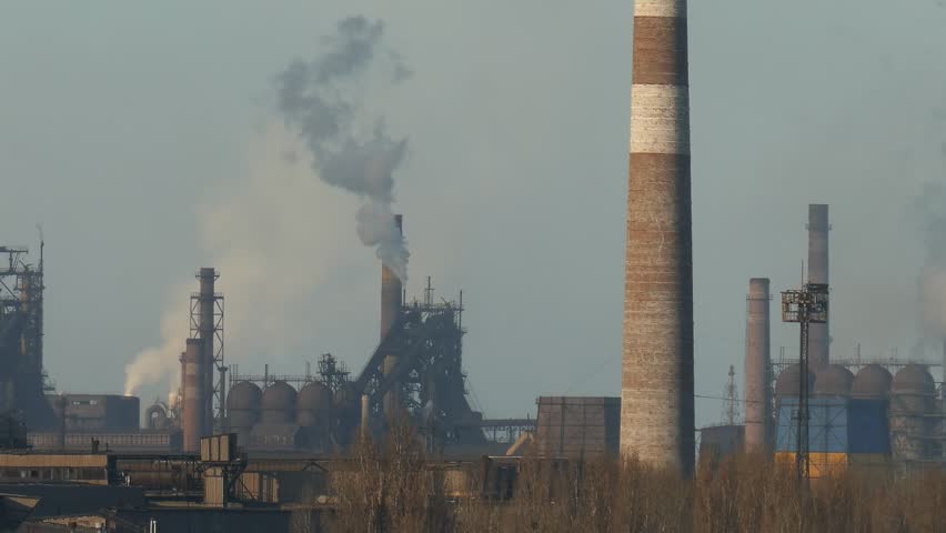 Wide panoramic shot of a large metallurgical or steel factory complex polluting the sky. Heavy industry structures with thick smoke coming out of chimneys and pipes. Environmental damage