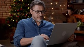 Older man working from home at Christmas in winter. Happy older white man with laptop at home, thinking, dark room with Christmas tree. - Powered by Shutterstock - Get 15% off with code: PIKWIZARD15