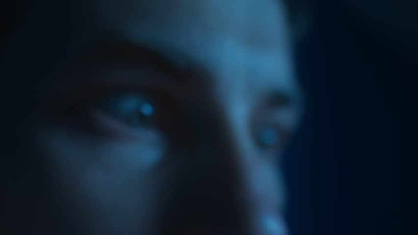 Man eyes with a crazy look nervously staring at the screen with a reflection of social media news feed at night. Harm of excessive content viewing, anxiety, digital addiction, doomscrolling.