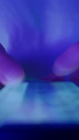 Close-up of hands holding a smartphone and quickly scrolling through social media feeds at night in neon light. Doom scrolling, phone overuse and digital addiction concept. Vertical video
