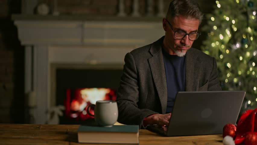 Happy smiling mid adult man using laptop computer at home, sitting at desk in Christmas decorated cosy room. Fireplace and Christmas tree in background. Businessman shopping online searching for gift. - Powered by Shutterstock - Get 15% off with code: PIKWIZARD15
