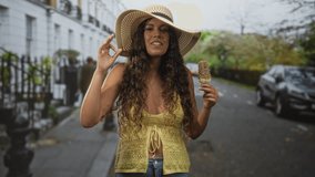 Hispanic woman holds a nut coated ice cream and makes an ok sign while smiling on an urban street wearing a straw hat and yellow top; carefree joy. - Powered by Shutterstock - Get 15% off with code: PIKWIZARD15