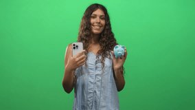 Teenage woman holds smartphone near chest and blue piggybank at shoulder in a studio green screen setting; financial confidence. - Powered by Shutterstock - Get 15% off with code: PIKWIZARD15