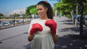 Woman in white tee wearing red boxing gloves holds fists up on urban street with wary smile and guarded stance; determination resilience. - Powered by Shutterstock - Get 15% off with code: PIKWIZARD15