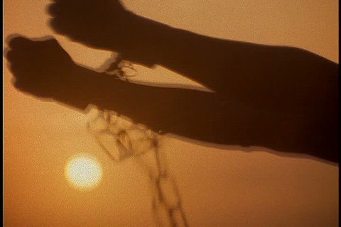 Slavery reenactment. CU silhouette of chained arms raised to the sun against a dark orange sky.