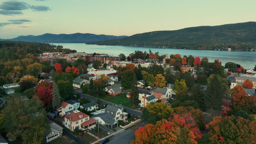Adirondack Mountain, New York State. Aerial view of a town nestled beside a lake during autumn, with trees displaying a mix of red, orange, and yellow foliage.