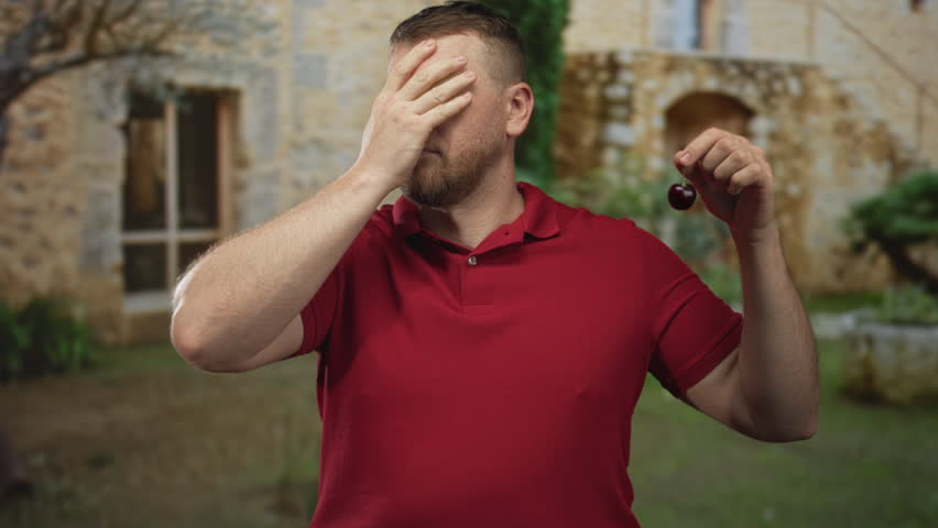 Man in red polo shirt holding cherry while hand covers left eye in stone building entrance; embarrassment confusion.