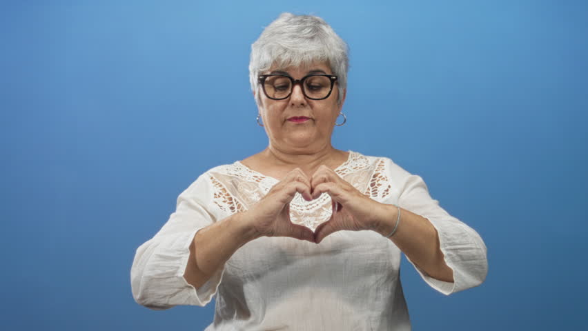 Senior woman makes heart with hands smiling, bare fingers visible, wearing white blouse and glasses in studio; warmth joy. - Powered by Shutterstock - Get 15% off with code: PIKWIZARD15