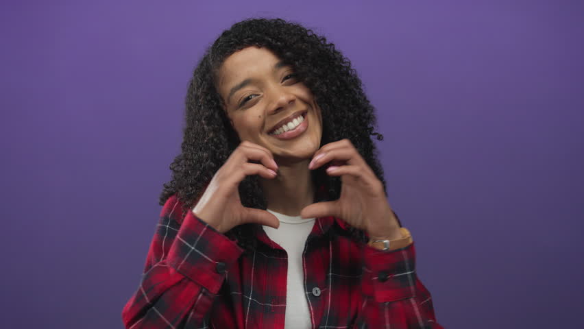 African american woman in red plaid shirt smiling forms heart gesture in purple studio; love warmth.