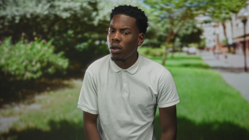 Man face looks surprised wearing white polo shirt amid green foliage on urban street; surprise curiosity.