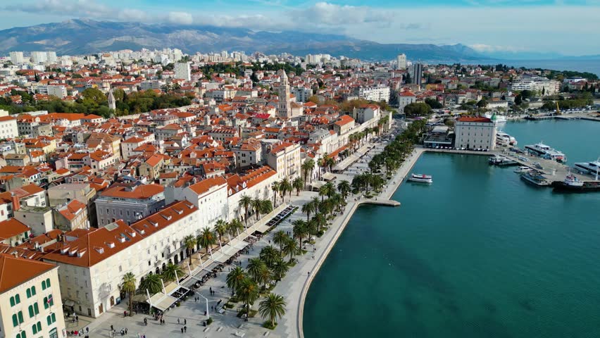 Aerial view of Split, Croatia, highlighting its historic red-tiled roofs, vibrant Riva promenade, and the stunning Adriatic Sea against a backdrop of mountains.