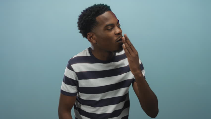 Man in striped shirt blowing kiss with hand near lips in studio with light blue backdrop; affection love.