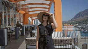 Woman holds brim of straw sunhat with hand, smiling and wearing sunglasses on cruise terrace building with orange canopy and striped chairs; vacation serenity. - Powered by Shutterstock - Get 15% off with code: PIKWIZARD15