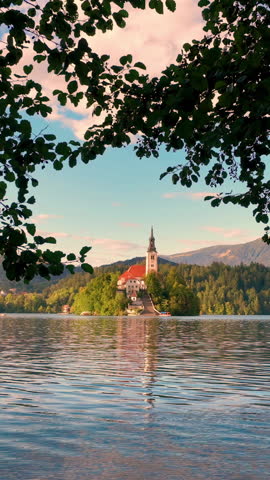 sunlit island chapel lake aerial sunset, drone pans over turquoise water surrounding small wooded islet, Bled