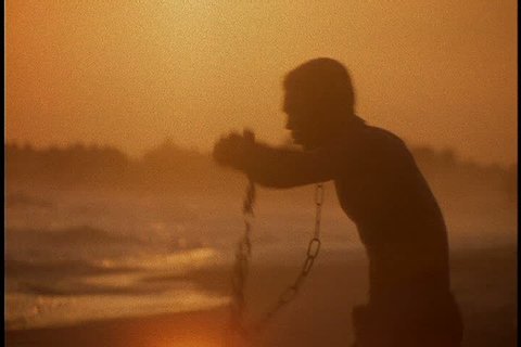 Slavery reenactment in Ghana, Africa. MS silhouette of black man in chains crouching on beach. He stands and raises his arms to the sun in a dark orange colored sky.