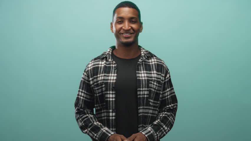 Young black man smiles with hands clasped and holds a small prop in studio teal backdrop, frontal framing; friendly confidence.