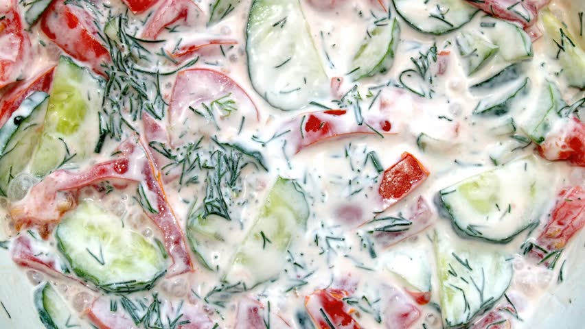 Close-up, full frame background of fresh vegetable salad with creamy sauce. Diced cucumbers, tomatoes, and dill mixed with sour cream or yogurt. Healthy summer food and culinary texture.