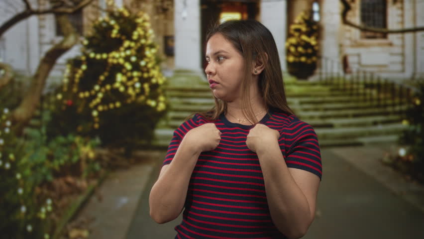 Woman points both hands to her chest in front of a building with holiday lights on steps; assertion defiance.