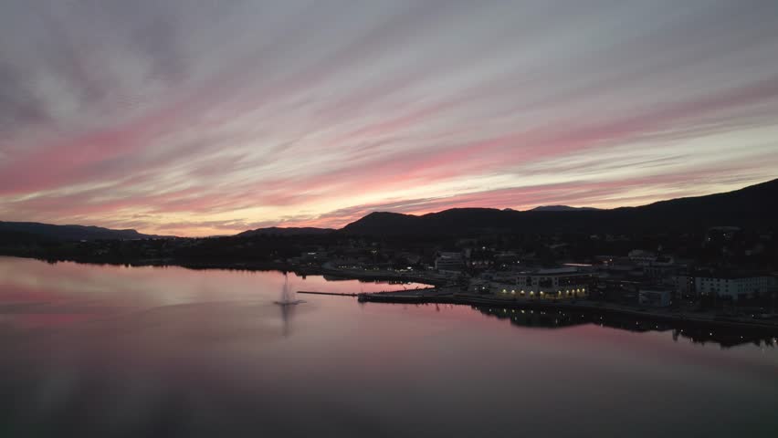 Atmospheric drone footage of a red-pink sunset over the fjord in Fauske. Still water reflects dramatic evening clouds and distant mountains. Peaceful, moody nordic scenery at dusk.