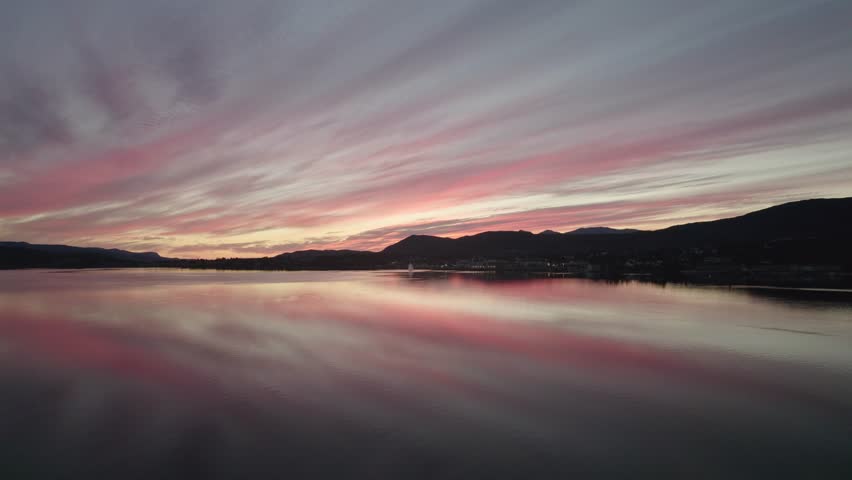 Soft pink and red sunset reflecting on calm water near Fauske, Norway. Smooth drone motion captures serene mountains and colorful clouds above a still fjord surface. Peaceful nordic evening landscape.