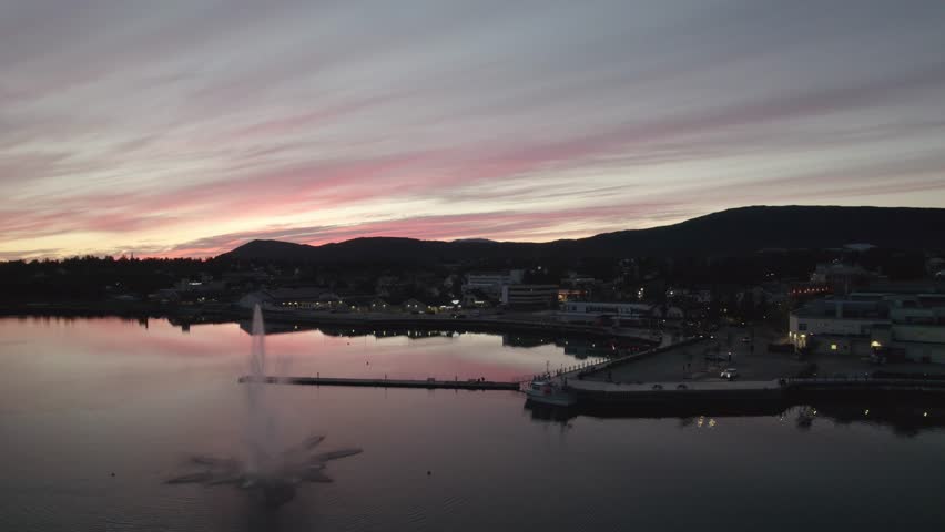 Vibrant red and pink clouds over the fjord at dusk in Fauske, Norway. Gentle drone flight over quiet water with soft reflections and dramatic mountain silhouettes. Tranquil arctic sunset mood.