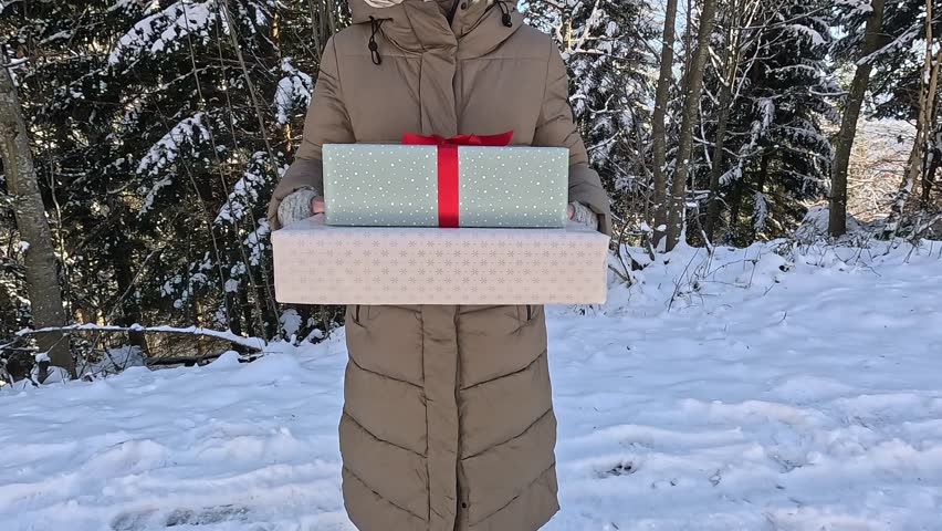 Woman in a warm winter coat holding two beautifully wrapped christmas presents while standing in a snowy forest. Festive season concept showing the joy of giving and holiday celebration