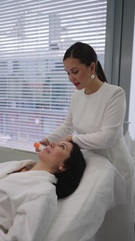 Vertical portrait of female cosmetologist performing rejuvenating ultrasound lifting facial treatment on adult woman face at modern aesthetic clinic, focusing on anti-aging and skin contouring.