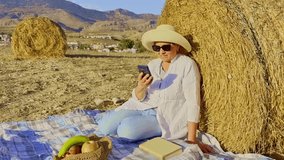 A senior woman sits on a soft haystack, enjoying a peaceful picnic outdoors. She holds a mobile phone in her hand, smiling and engaging in a pleasant conversation. Fresh fruits are laid out . - Powered by Shutterstock - Get 15% off with code: PIKWIZARD15