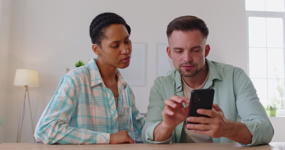 Happy excited couple using smartphone to read news about lottery win at home. Young amazed man and woman reading information on phone screen, winners with surprised expression on faces give high five