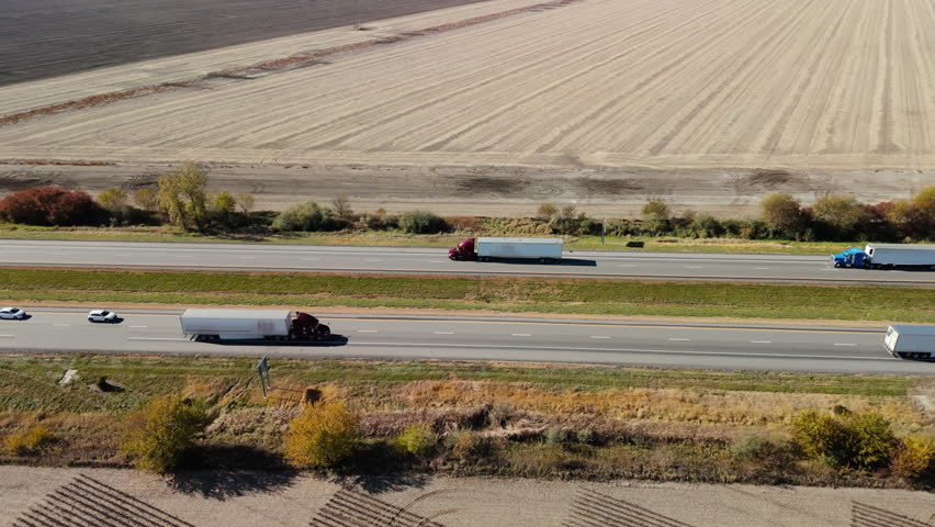 Multiple semi-trucks and cars traveling along a busy highway surrounded by flat agricultural fields. The clip shows long-haul transportation, logistics, and supply chain