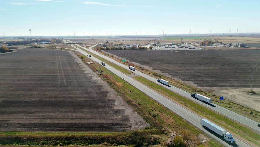 Multiple semi-trucks and cars traveling forward on a busy highway surrounded by flat plowed agricultural fields. The clip shows long-haul transportation, logistics, and supply chain